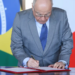 Autoridade masculina de aproximadamente 60-70 anos, vestindo terno cinza e gravata azul estampada, assina documento oficial à mesa.
