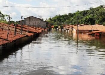 Casas com telhados vermelhos submersas por enchente em rua alagada, céu parcialmente nublado