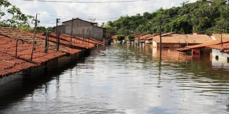 Casas com telhados vermelhos submersas por enchente em rua alagada, céu parcialmente nublado