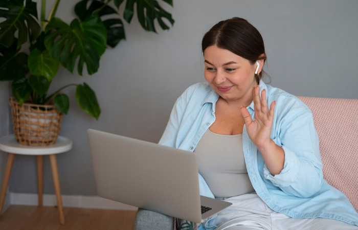 Mulher sorridente acena para laptop usando camiseta cinza e camisa azul clara, sentada com fone de ouvido sem fio