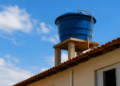 Caixa d’água azul apoiada em estrutura de concreto sobre telhado de casa com céu azul ao fundo