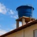 Caixa d’água azul apoiada em estrutura de concreto sobre telhado de casa com céu azul ao fundo
