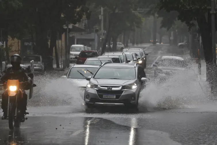 100 mm de chuva por dia e ventos de até 100 km/h no RS: alerta laranja do Inmet prevê granizo e danos 2 Pessoas caminhando sob chuva forte com guarda-chuvas e capas plásticas em calçada movimentada, carros ao fundo