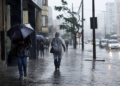Pessoas caminhando sob chuva forte com guarda-chuvas e capas plásticas em calçada movimentada, carros ao fundo
