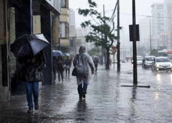 Pessoas caminhando sob chuva forte com guarda-chuvas e capas plásticas em calçada movimentada, carros ao fundo