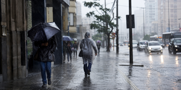 Pessoas caminhando sob chuva forte com guarda-chuvas e capas plásticas em calçada movimentada, carros ao fundo