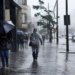 Pessoas caminhando sob chuva forte com guarda-chuvas e capas plásticas em calçada movimentada, carros ao fundo
