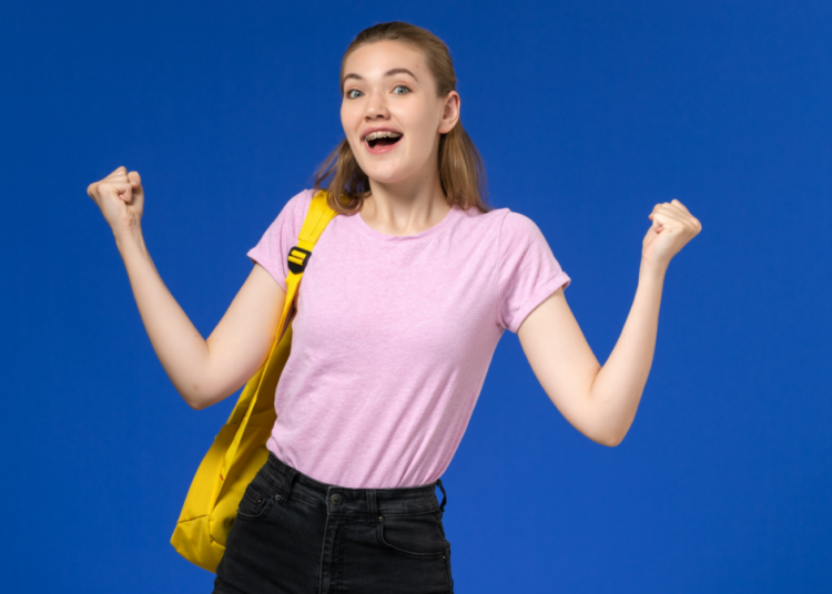Adolescente sorridente comemora conquista com mochila amarela em fundo azul.