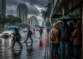 Pessoas com guarda-chuvas atravessam avenida alagada enquanto grupo se abriga na calçada sob chuva intensa