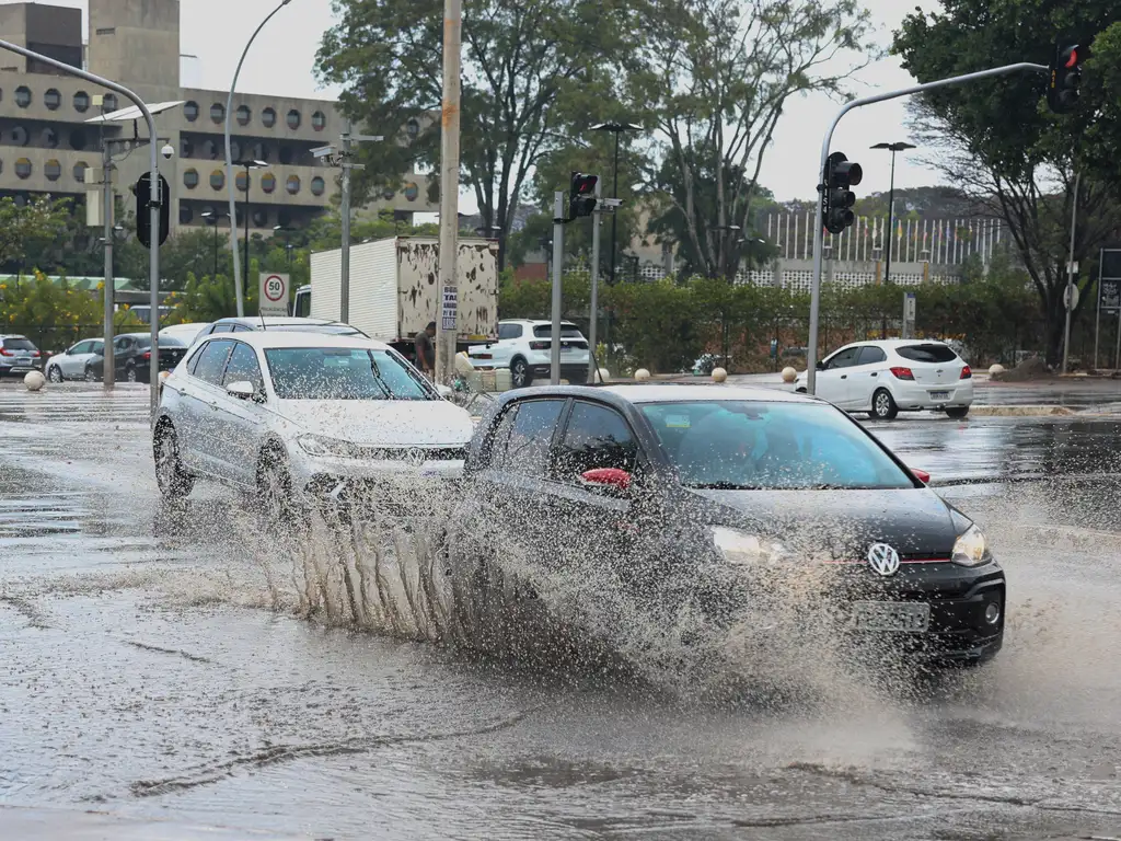 Carros atravessam rua alagada formando grande onda de água em cruzamento urbano