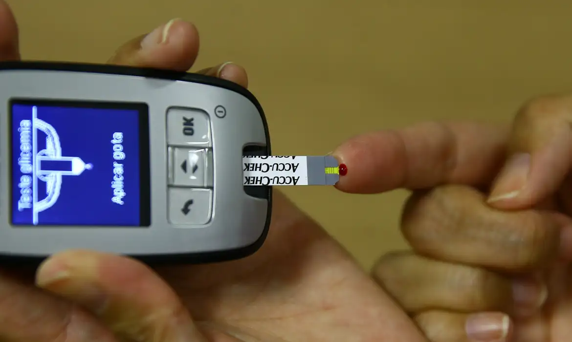 Pessoa realiza teste de glicemia usando aparelho ACCU-CHEK e fita com gota de sangue no dedo