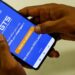 Mãos masculinas seguram celular exibindo tela do app FGTS da Caixa, botão laranja em destaque