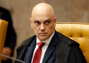Alexandre de Moraes sentado em cadeira amarela no STF, usando terno e gravata vermelha, expressão séria.