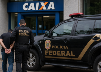 agente da Polícia Federal conduz homem algemado próximo a viatura preta diante de agência da Caixa