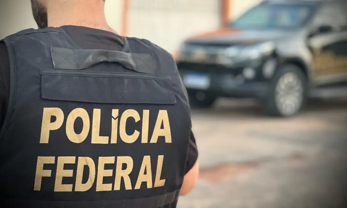 R$ 45 milhões desviados de correntistas e beneficiários da Caixa: PF investiga fraude no RJ e SP 2 Agente da Polícia Federal de colete preto, visto de costas, próximo a viatura escura desfocada ao fundo
