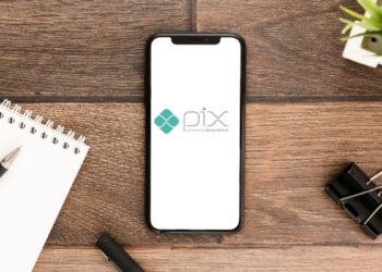 Smartphone exibindo logomarca do PIX no centro de mesa de madeira com caderno, caneta e clipe.