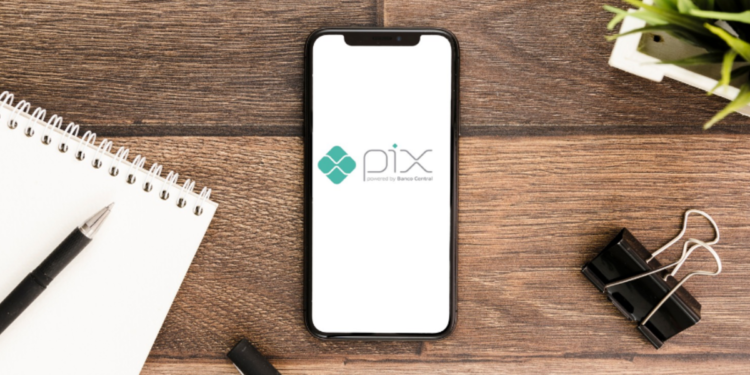 Smartphone exibindo logomarca do PIX no centro de mesa de madeira com caderno, caneta e clipe.