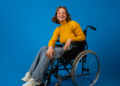 Jovem cadeirante sorridente com suéter amarelo em fundo azul.