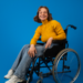 Jovem cadeirante sorridente com suéter amarelo em fundo azul.