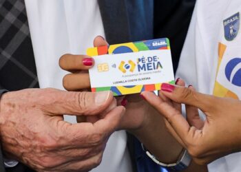 Mãos segurando cartão colorido Pé-de-Meia com nome de estudante e brasão do Brasil à mostra