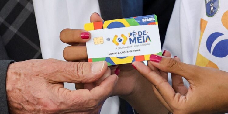 Mãos segurando cartão colorido Pé-de-Meia com nome de estudante e brasão do Brasil à mostra