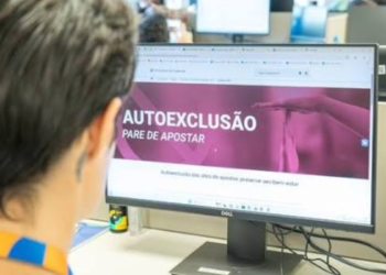 Pessoa olhando monitor com tela de autoexclusão e mensagem “pare de apostar” em escritório.
