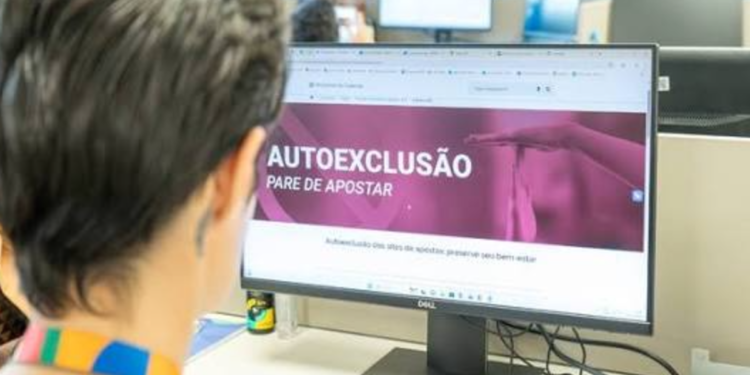 Pessoa olhando monitor com tela de autoexclusão e mensagem “pare de apostar” em escritório.