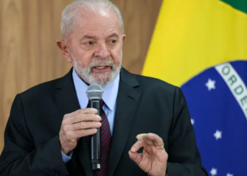 Lula segurando microfone em frente à bandeira brasileira, fazendo gesto com a mão.