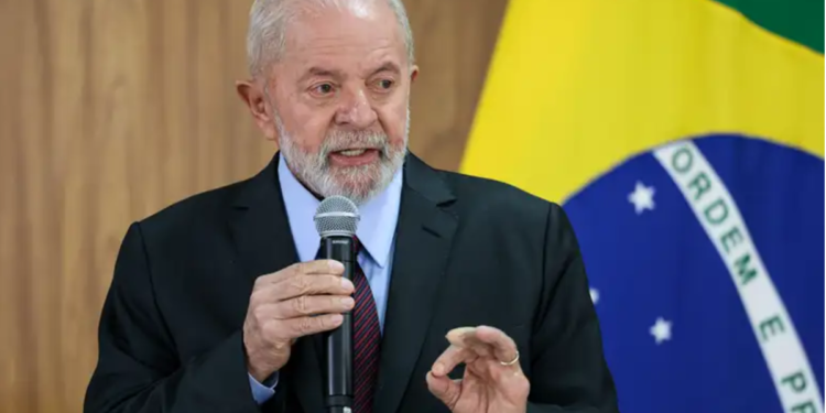 Lula segurando microfone em frente à bandeira brasileira, fazendo gesto com a mão.