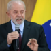 Lula segurando microfone em frente à bandeira brasileira, fazendo gesto com a mão.