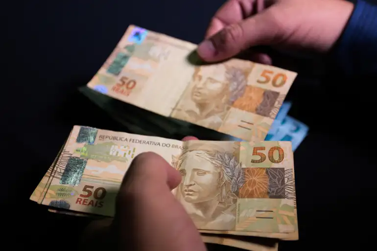 R$ 547 milhões para ressarcir beneficiários do INSS: vítimas de descontos indevidos receberão valores 2 Mãos de pessoa contando várias notas de 50 reais com fundo preto e detalhamento das cédulas