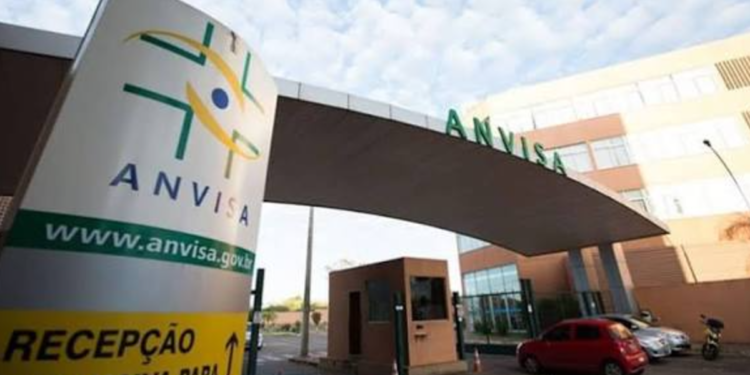 Entrada da sede da Anvisa com placa, estrutura moderna, carros e céu azul ao fundo.
