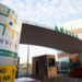 Entrada da sede da Anvisa com placa, estrutura moderna, carros e céu azul ao fundo.