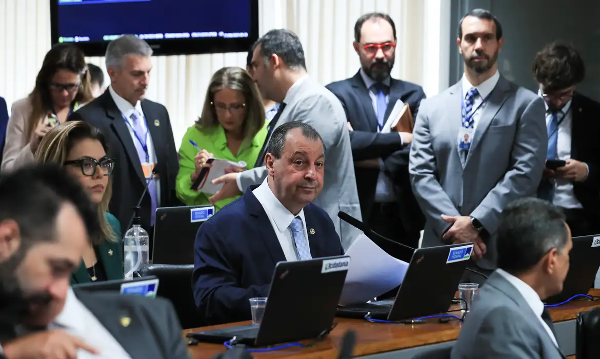 Senadores reunidos em sessão, homens e mulheres em terno, computadores, crachás e papéis sobre a mesa