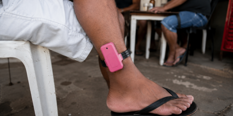 Homem com tornozeleira eletrônica rosa sentado de bermuda e chinelo em bar simples