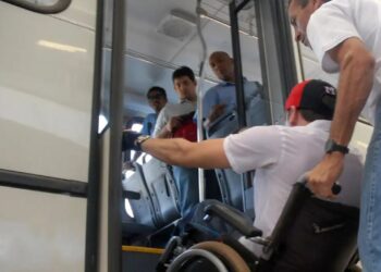 Homem em cadeira de rodas tentando entrar em ônibus com ajuda, observado por três passageiros no interior