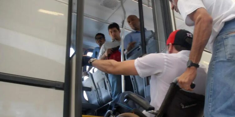 Homem em cadeira de rodas tentando entrar em ônibus com ajuda, observado por três passageiros no interior