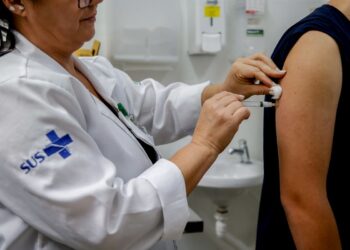 Profissional de saúde do SUS aplicando vacina no braço de paciente com seringa e algodão