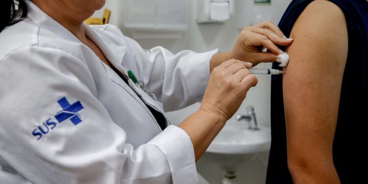 Profissional de saúde do SUS aplicando vacina no braço de paciente com seringa e algodão