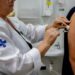 Profissional de saúde do SUS aplicando vacina no braço de paciente com seringa e algodão