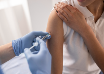 Mulher recebe vacina em consultório de saúde com profissional usando luvas.