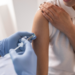 Mulher recebe vacina em consultório de saúde com profissional usando luvas.