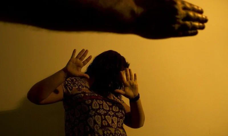 Mulher assustada cobre o rosto com as mãos enquanto braço ergue-se ameaçadoramente em fundo amarelo