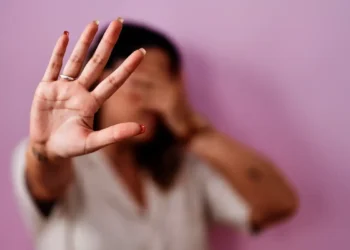 Mulher com a mão aberta à frente do rosto, palma à mostra, expressão de recusa contra fundo lilás