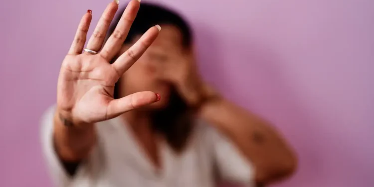Mulher com a mão aberta à frente do rosto, palma à mostra, expressão de recusa contra fundo lilás