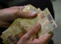 Mãos de uma pessoa contando cédulas de 50 reais sobre fundo escuro, em imagem que representa o crédito do último lote do abono salarial PIS/Pasep do calendário 2026, pago em 17 de agosto aos trabalhadores nascidos em novembro e dezembro, com valores proporcionais aos meses trabalhados em 2024