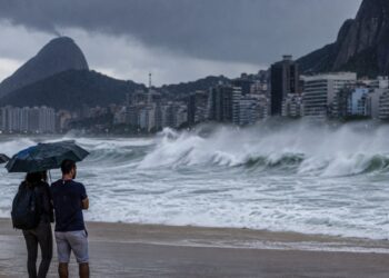 Ondas de até 3 metros atingem a orla do Rio de Janeiro durante o aviso de ressaca da Marinha, com pessoas de guarda-chuva observando o mar sob céu de chuva
