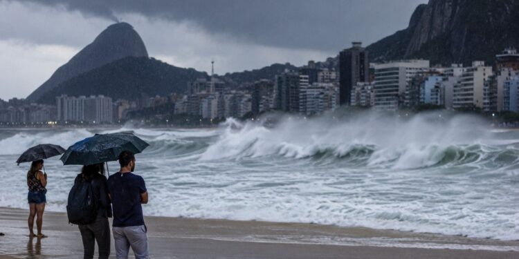 Ondas de até 3 metros atingem a orla do Rio de Janeiro durante o aviso de ressaca da Marinha, com pessoas de guarda-chuva observando o mar sob céu de chuva