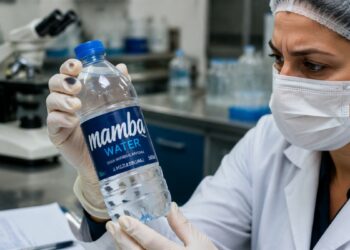 Profissional de laboratório com jaleco, máscara e luvas examina uma garrafa de água mineral Mamba Water, marca que teve lotes recolhidos pela Anvisa após contaminação por bactéria.