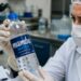 Profissional de laboratório com jaleco, máscara e luvas examina uma garrafa de água mineral Mamba Water, marca que teve lotes recolhidos pela Anvisa após contaminação por bactéria.